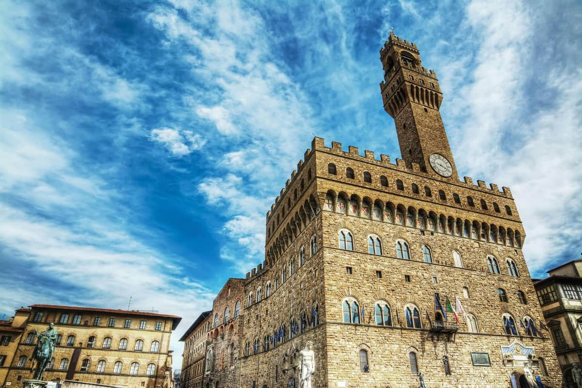 Historic stone building with arched windows and a tall clock tower under a partly cloudy blue sky. Statues line the building's facade.