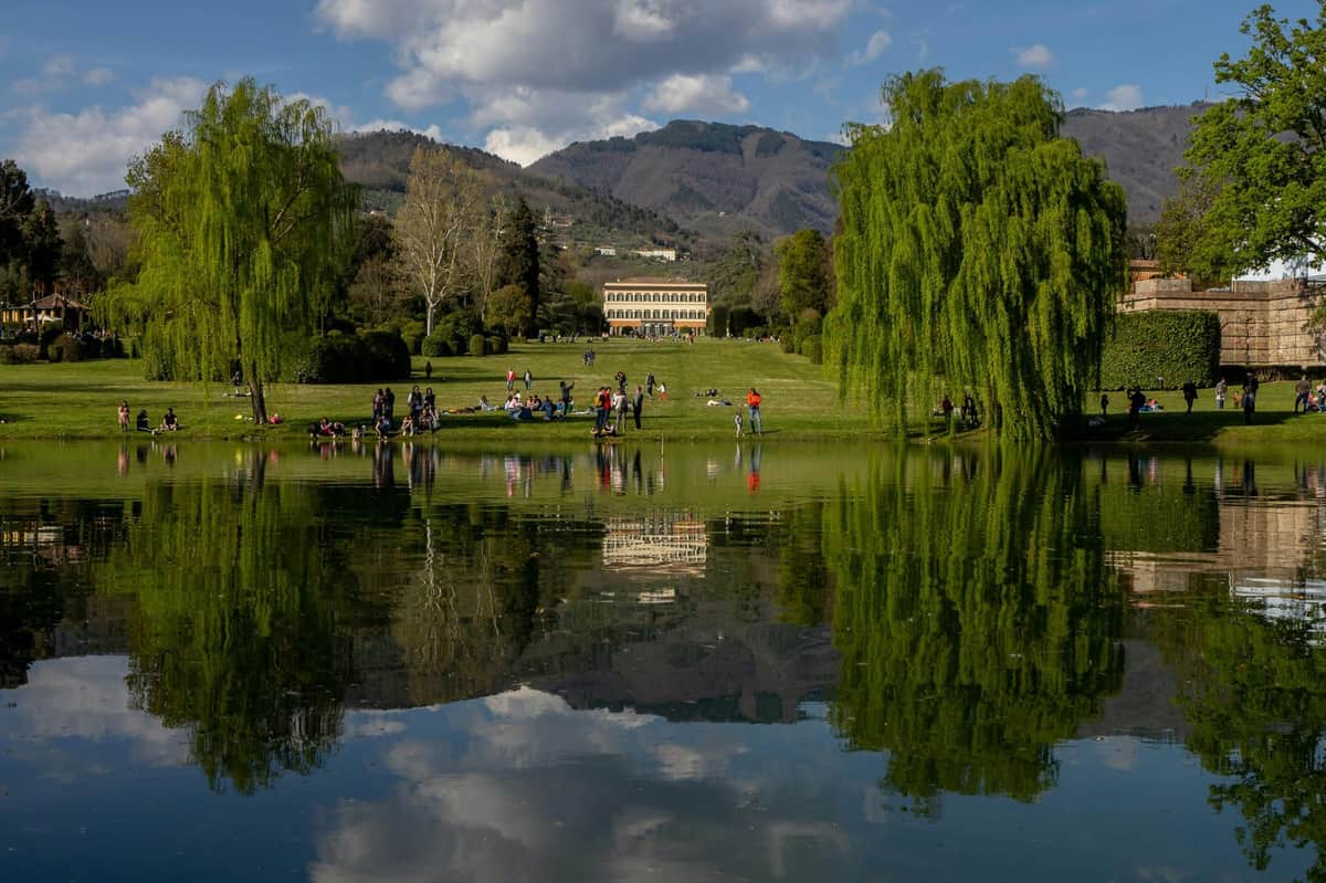A serene park with a reflective pond, lush greenery, people relaxing, and a building in the background under a partly cloudy sky.