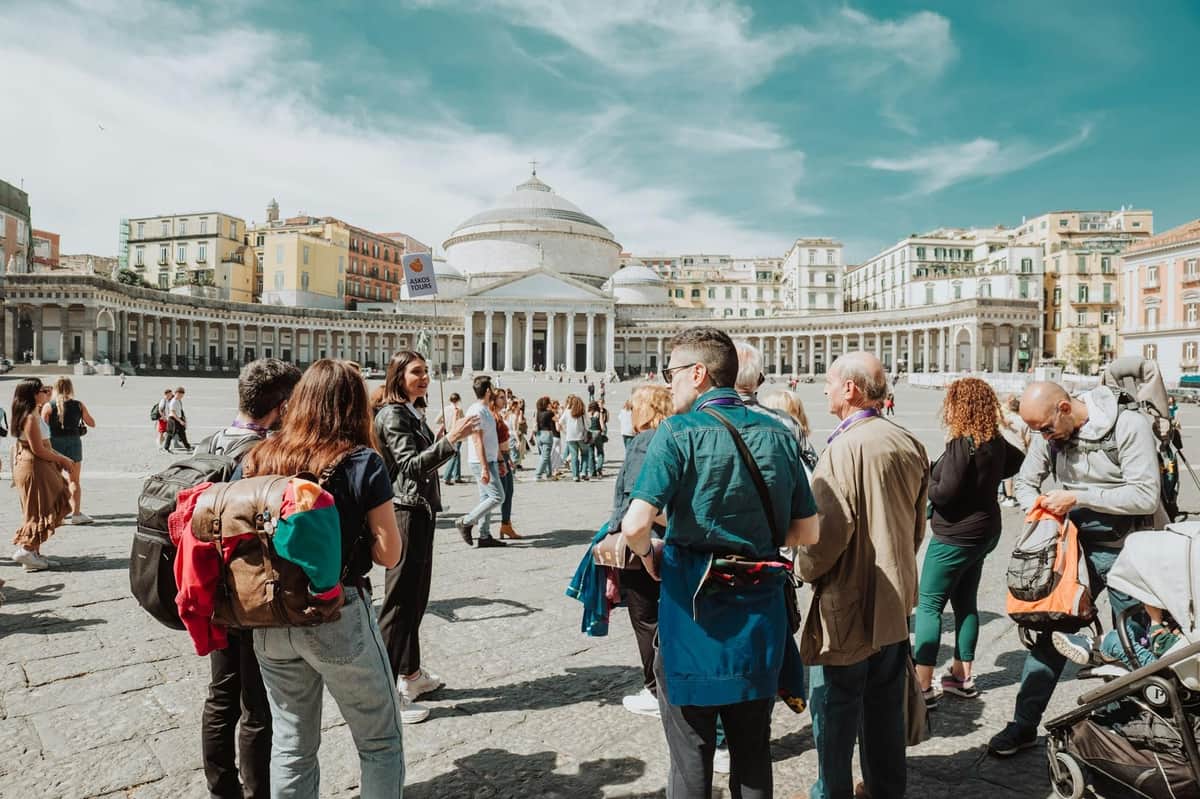 A group of people gathered in a square with a domed building and colonnades in the background under a blue sky.