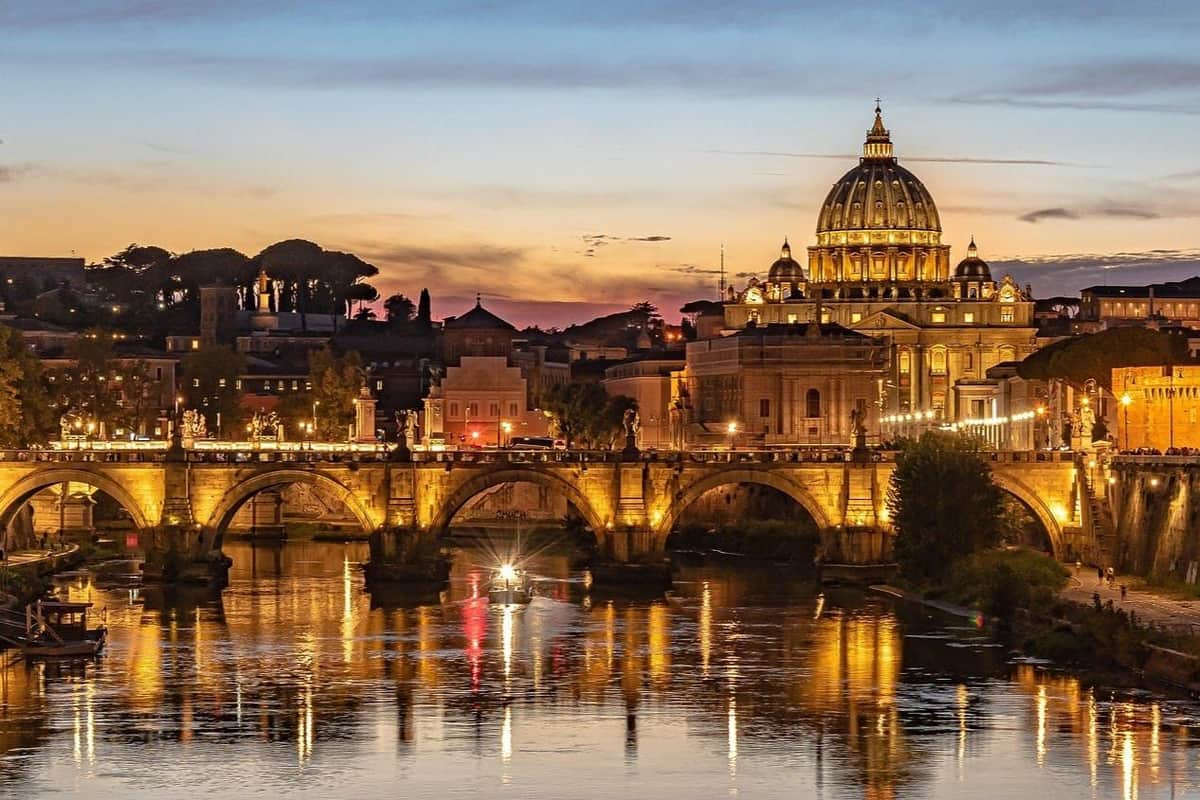 A bridge and illuminated buildings, including a large dome, reflected in a river at sunset with a boat passing underneath.