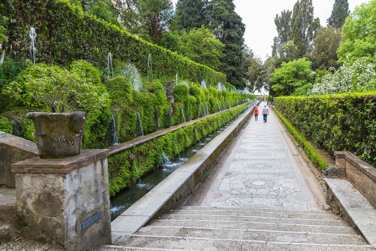 Villa D’Este and Villa Adriana: Skip the Line + Roundtrip from Rome Two people walking along a pathway with cascading water fountains and lush greenery on the left side.