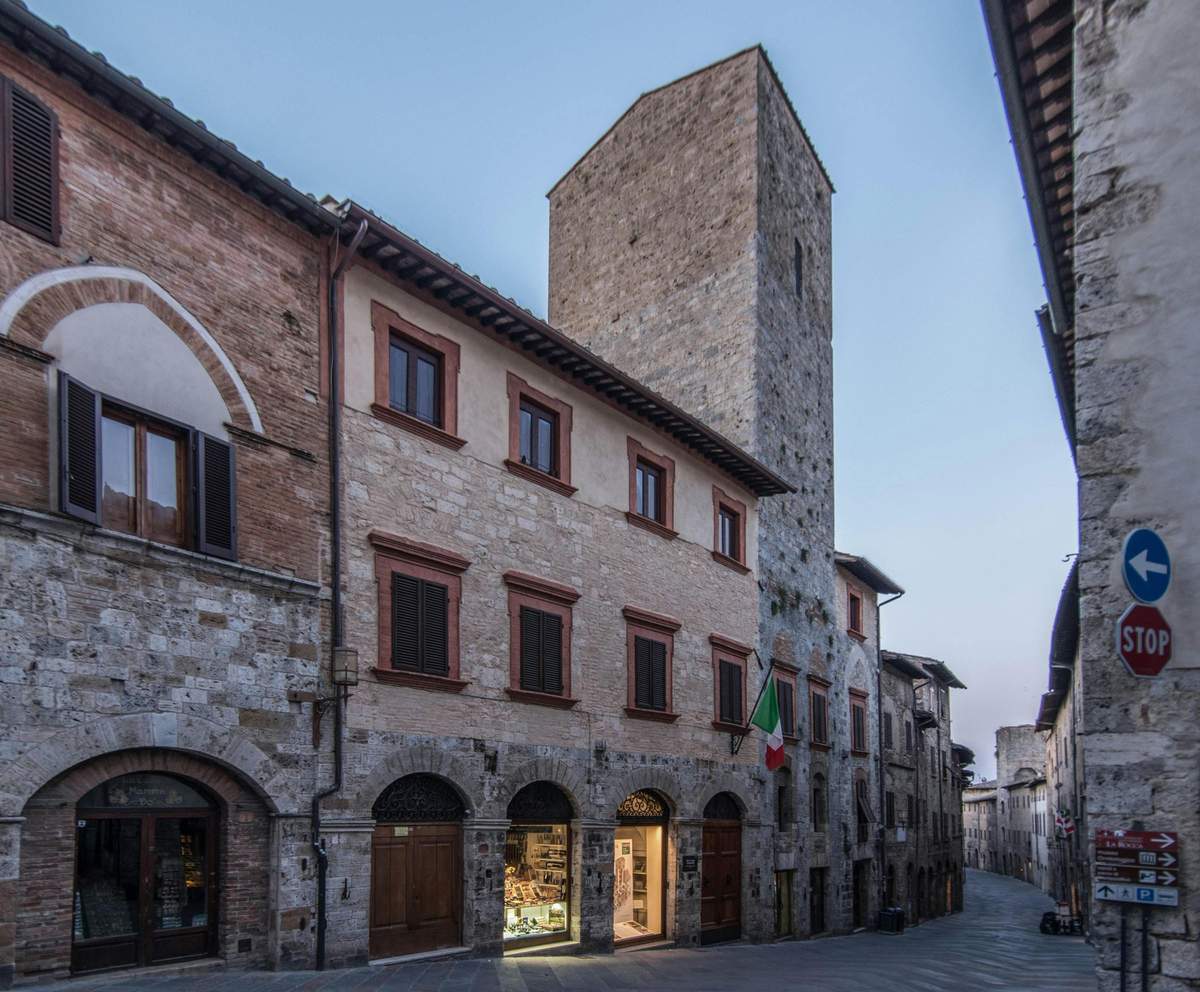 Historic stone building with a tall tower, featuring arched doorways, wooden shutters, and an Italian flag. Evening sky above.
