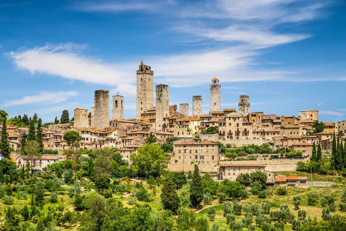 Pase San Gimignano Una ciudad histórica con numerosas y altas torres de piedra, rodeada de exuberante vegetación bajo un cielo azul con nubes dispersas.