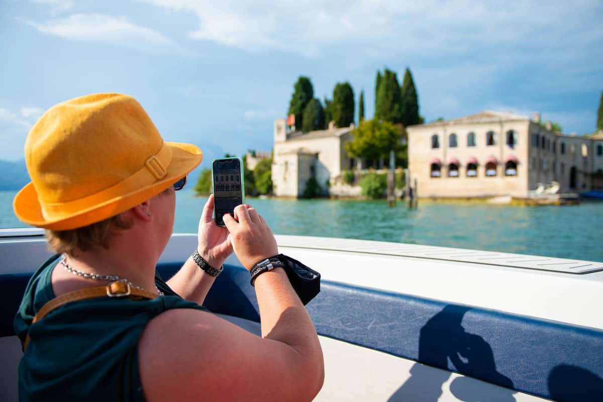 Lake Garda: 4-Hr Boat Tour with Scaliger Castles + Wine Tasting from Sirmione Person in a yellow hat taking a photo with a smartphone from a boat, capturing buildings and trees across a body of water.
