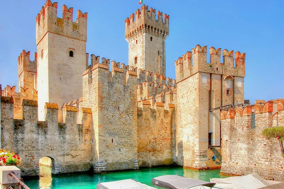 A medieval castle with crenelated walls and a tower, surrounded by a moat with boats, under a clear blue sky.