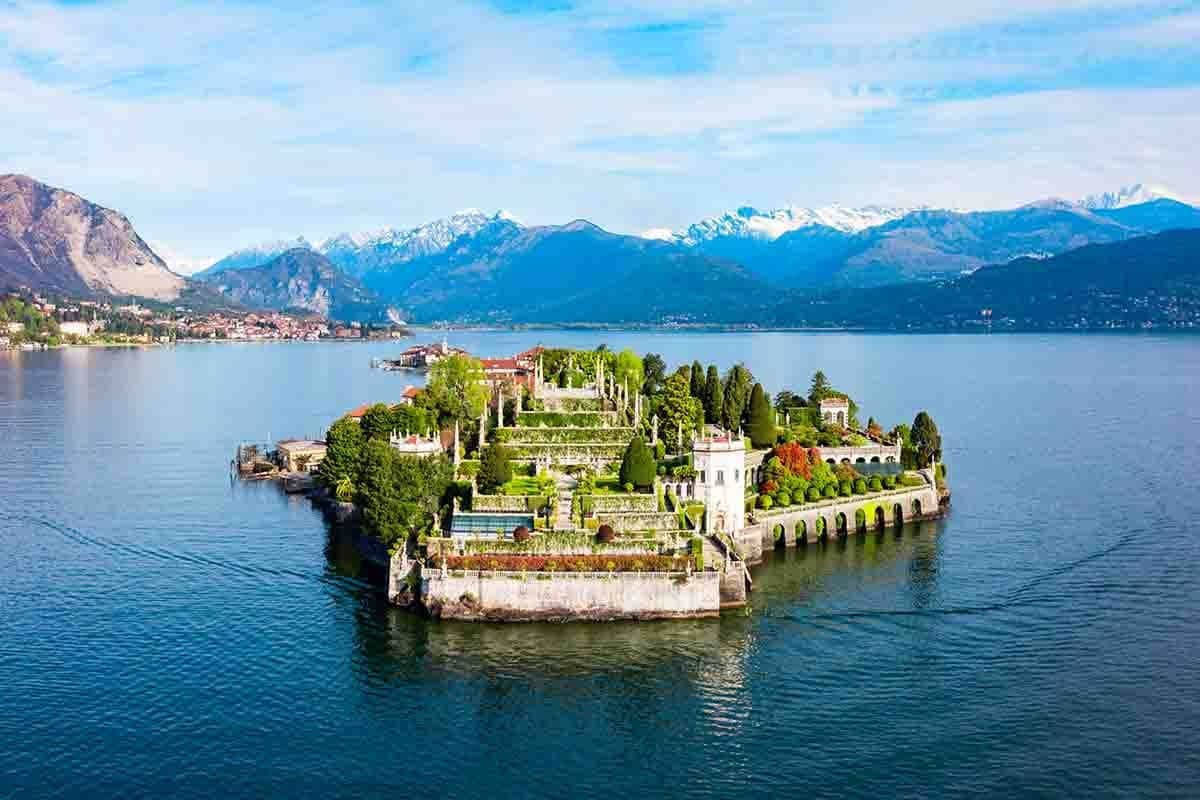 Lush, terraced garden on a small island surrounded by a lake, backed by snow-capped mountains under a partly cloudy sky.