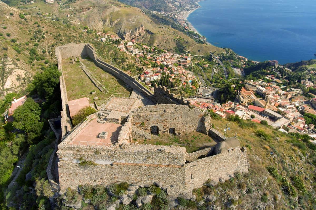 Castello di Taormina: Skip The Line Ticket + Audio Guide Aerial view of a stone fortress ruin on a hill overlooking a coastal town with winding streets and buildings near a blue sea.