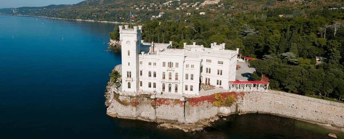 White stone castle situated on the edge of a cliff by a lake, surrounded by dense forest and hills.