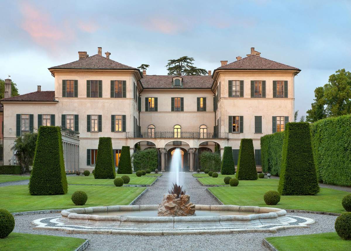 Large, elegant mansion with symmetrical towers, surrounded by manicured gardens, topiary trees, and a central fountain.