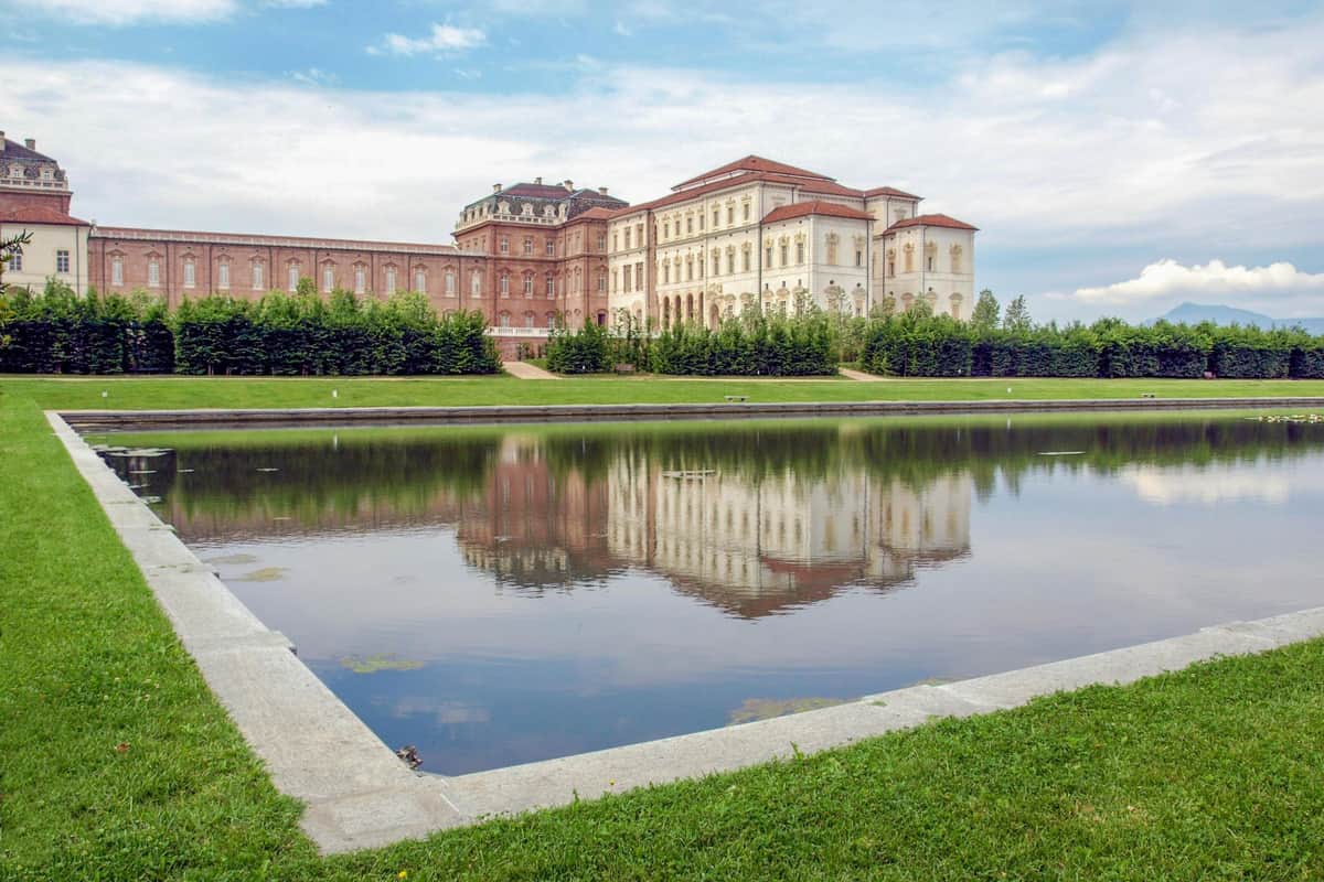 A large, historic building is reflected in a calm pond, with lush greenery and a manicured lawn under a partly cloudy sky.