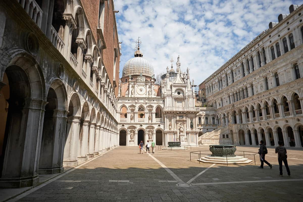 Doge's Palace: Fast Track Entry Three people walk in a large, ornate courtyard with domed and arched architecture under a partly cloudy sky.