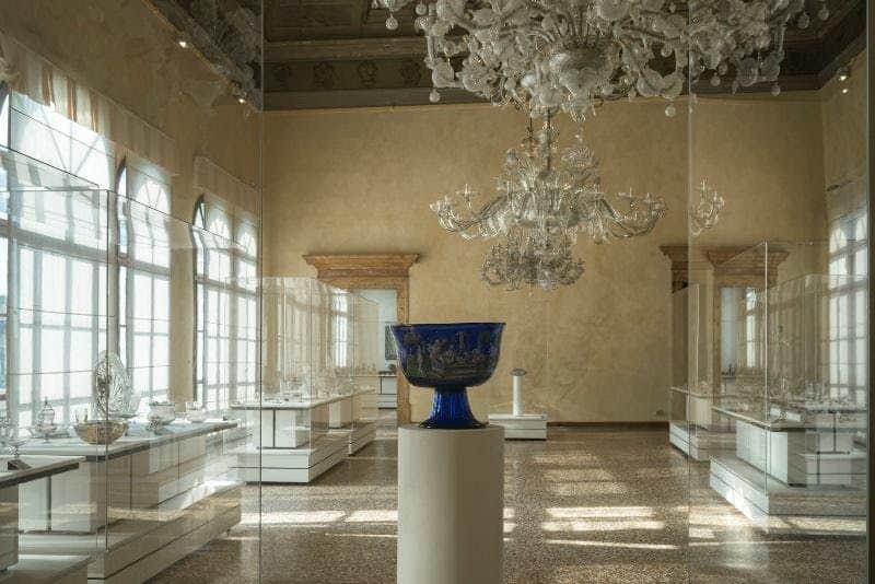 A blue glass bowl on a pedestal in a museum room with large windows, glass displays, and an ornate chandelier overhead.