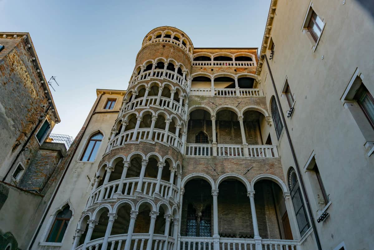 A multilevel spiral staircase with arched windows and a brick façade in a historic building against a clear sky.