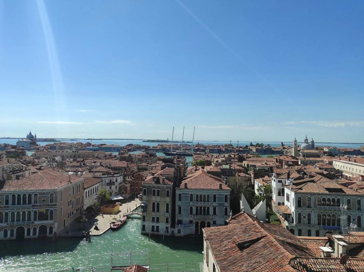 Elevated view of a city with red-tiled rooftops, canals, boats, and historic buildings under a clear blue sky.