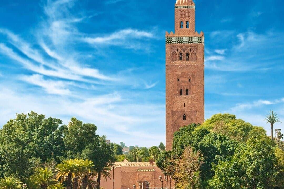 Tall brick minaret tower, surrounded by lush greenery, under a bright blue sky with scattered white clouds.