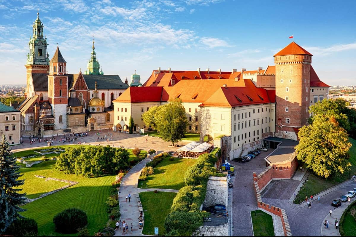 A historic European castle complex with red-roofed buildings, a church, and a courtyard surrounded by greenery under a blue sky.
