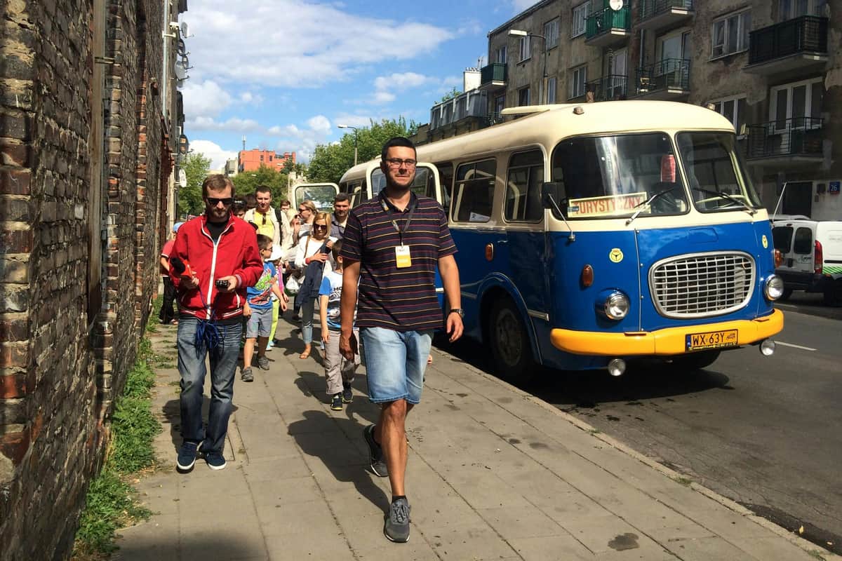 Retro Bus Tour Dark Side of Warsaw A group of people walks on a sidewalk next to a blue and cream bus, with buildings and a brick wall in the background.