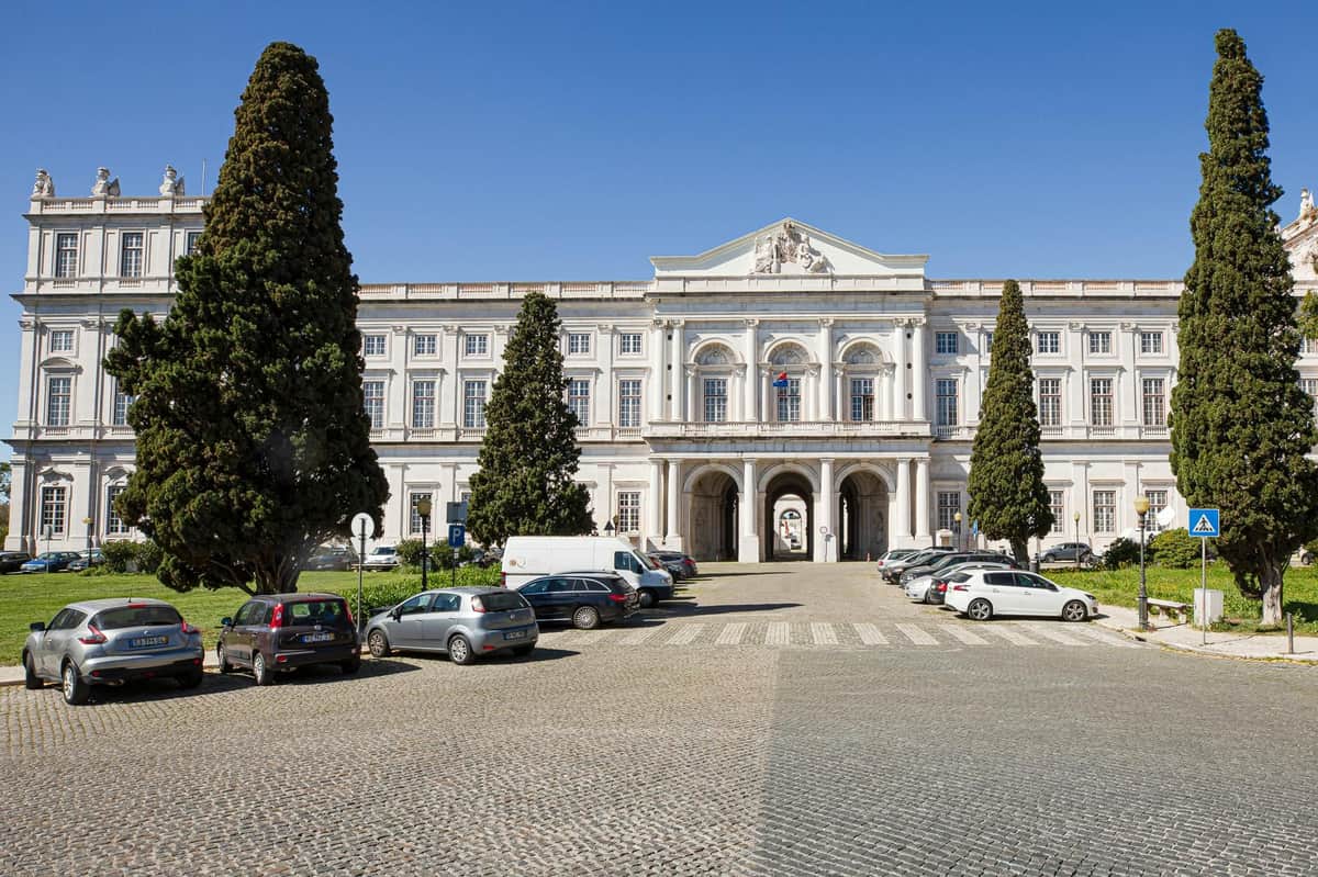 Large neoclassical building with arched entrance, flanked by tall trees, parked cars on cobblestone driveway, under clear blue sky.