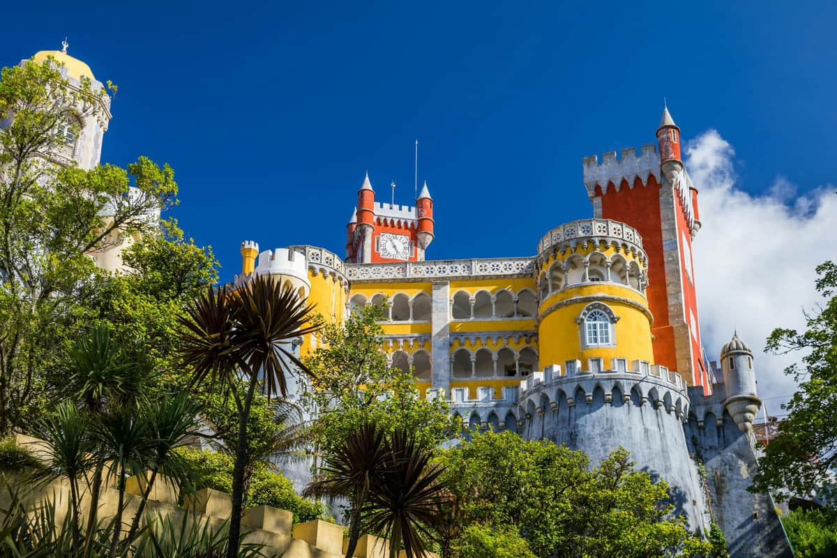 Un castillo de colores, con paredes amarillas, rojas y blancas, rodeado de árboles bajo un cielo azul y despejado.