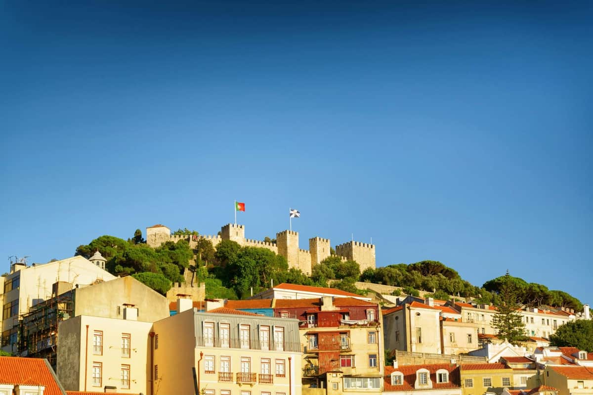 A castle with flags atop a hill, surrounded by greenery, with colorful buildings in the foreground under a clear blue sky.