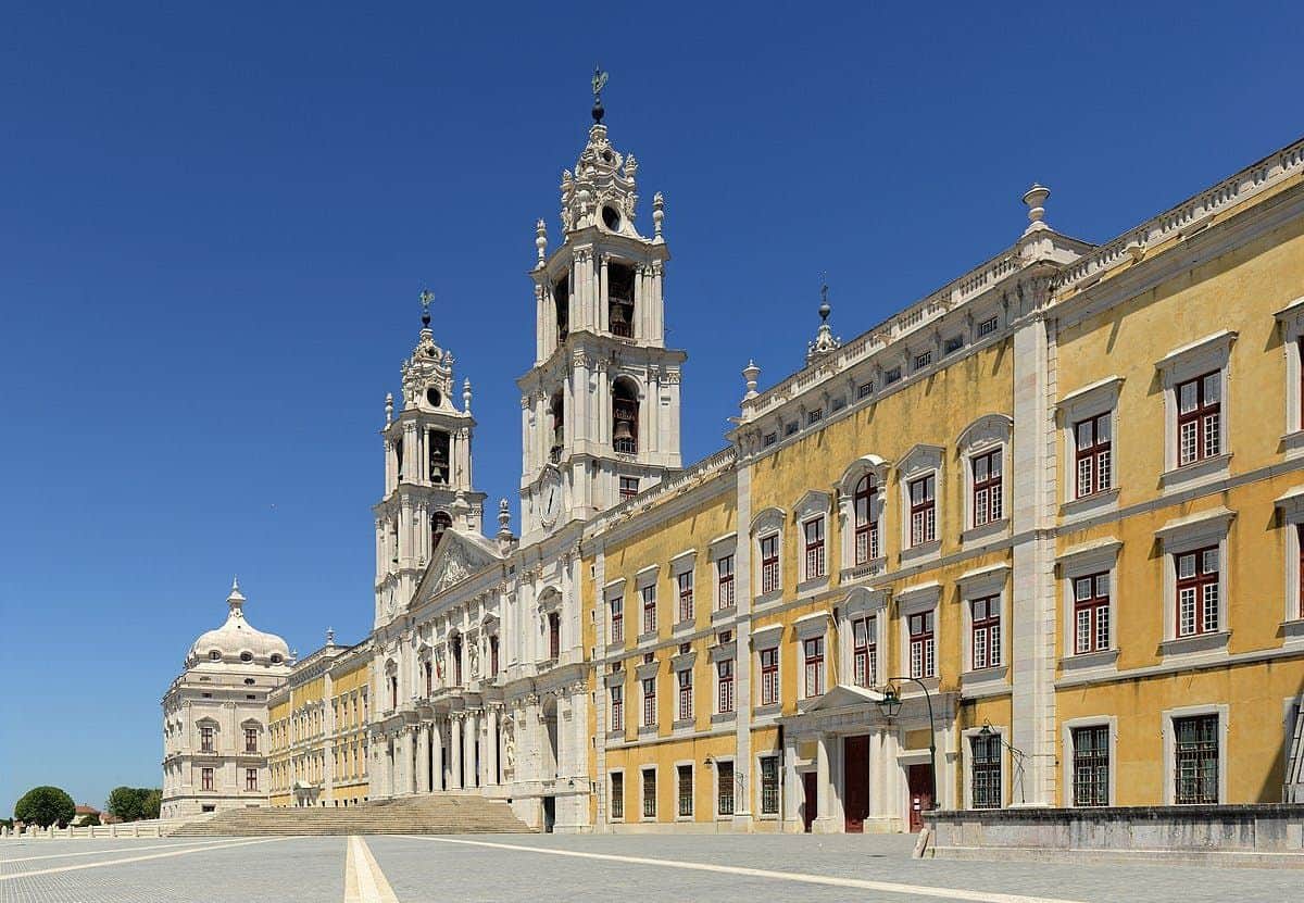 A large yellow and white baroque building with two tall towers and numerous windows under a clear blue sky.