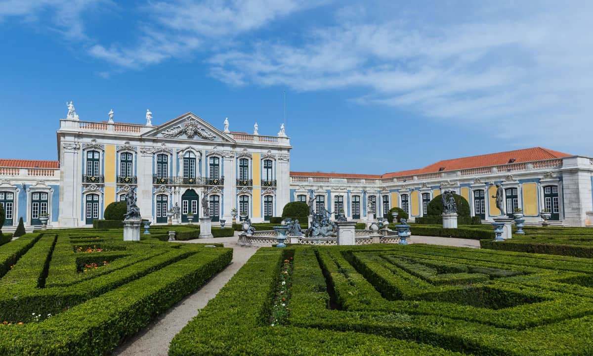 Elegant palace with ornate architecture, manicured garden with hedges, statues, and pathways under a clear blue sky.