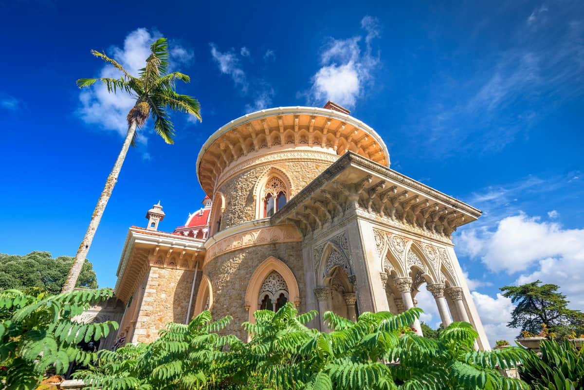 Ornate stone building with arched windows, surrounded by lush greenery, under a bright blue sky with scattered clouds.