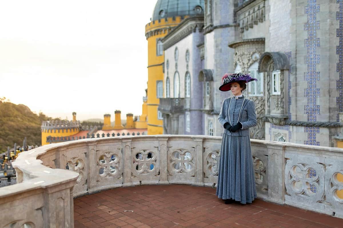 A person dressed in historical attire stands on a tiled balcony with ornate railings, with a castle in the background.