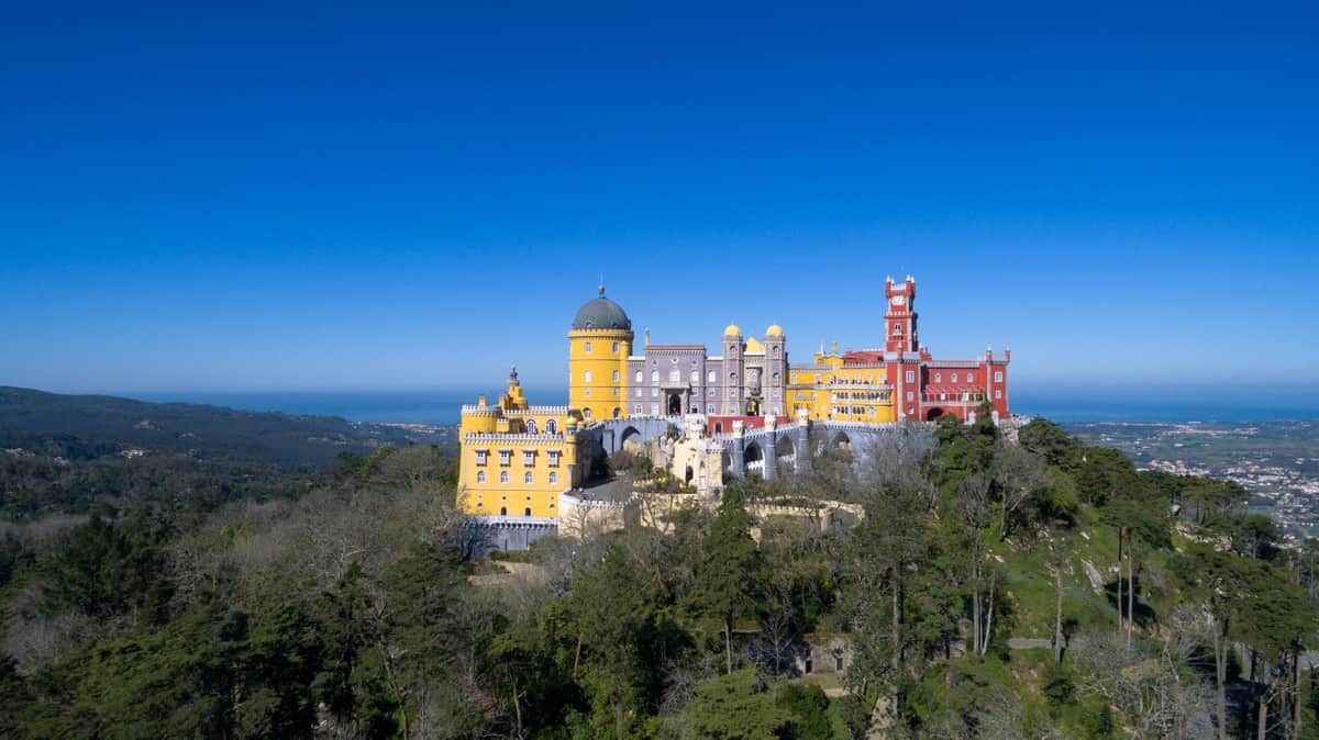 Colorful palace with yellow, purple, and red sections on a hilltop surrounded by trees, set under a clear blue sky.