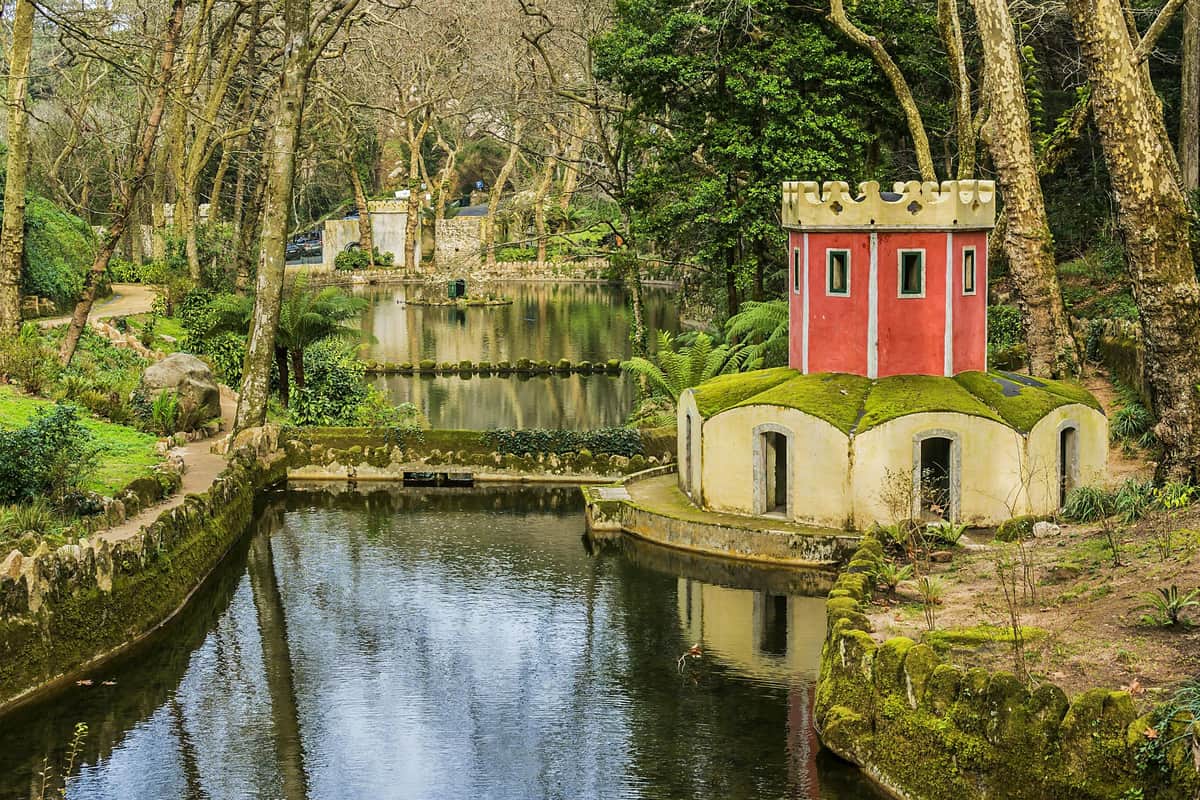 A small, red and yellow castle-like structure beside a reflective pond surrounded by trees and greenery in a park.