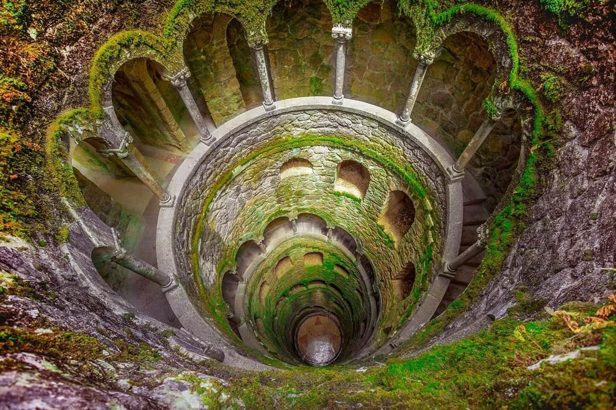 A stone spiral staircase with arches and moss-covered walls descends into a deep pit.