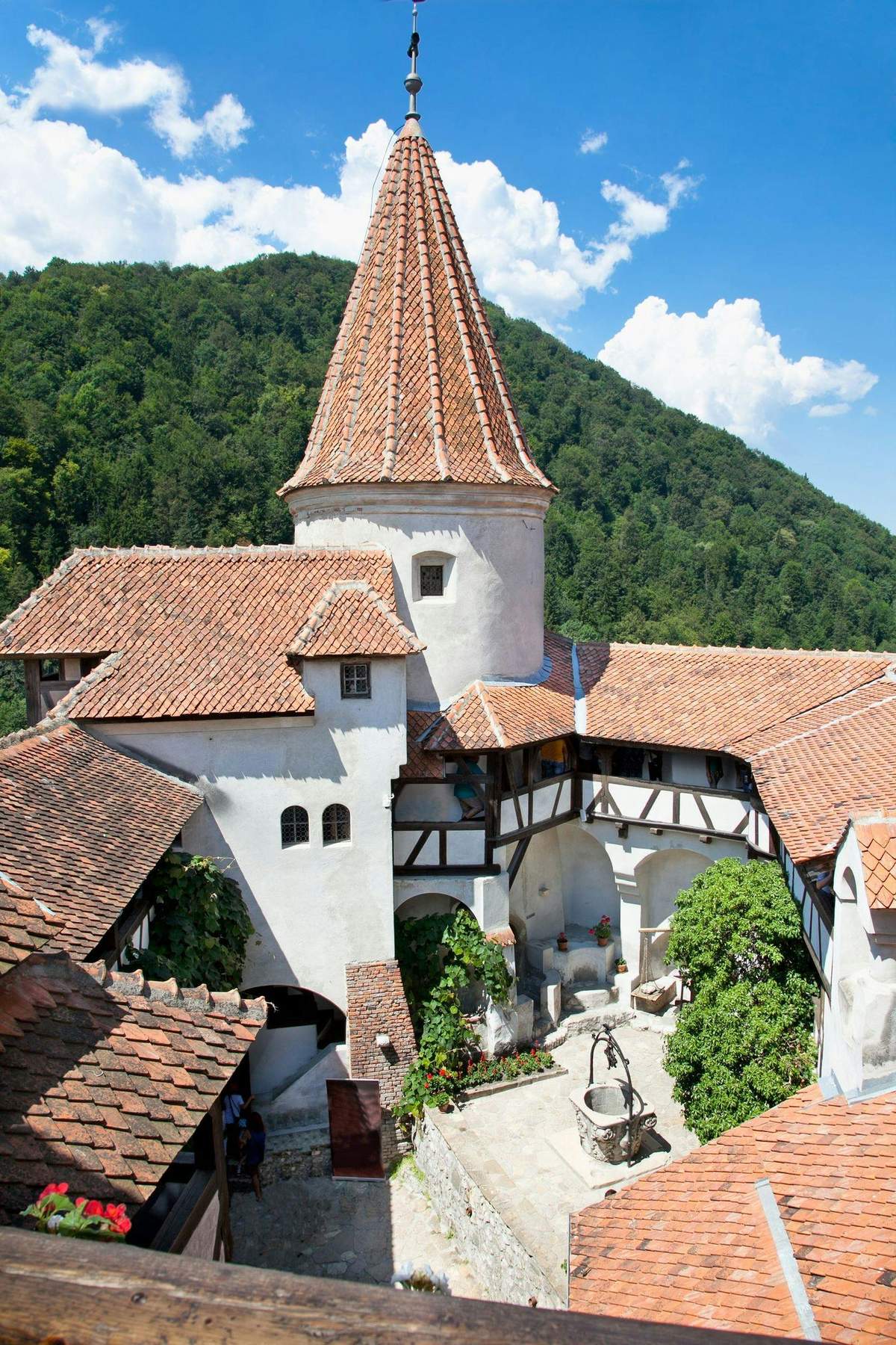 Medieval castle courtyard with a well, surrounded by buildings with red-tiled roofs and nestled against a forested hillside.
