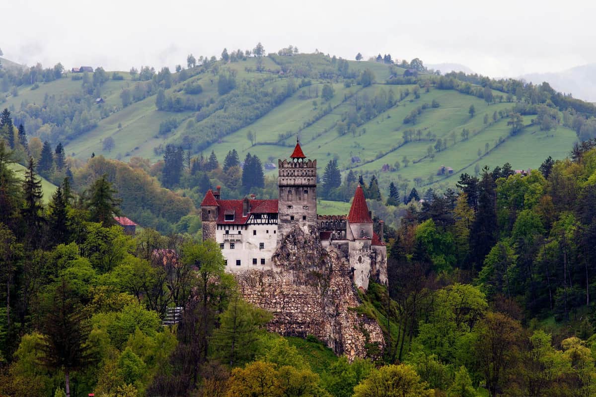 Castle with red rooftops situated atop a rocky hill, surrounded by green forests and rolling hills in the background.