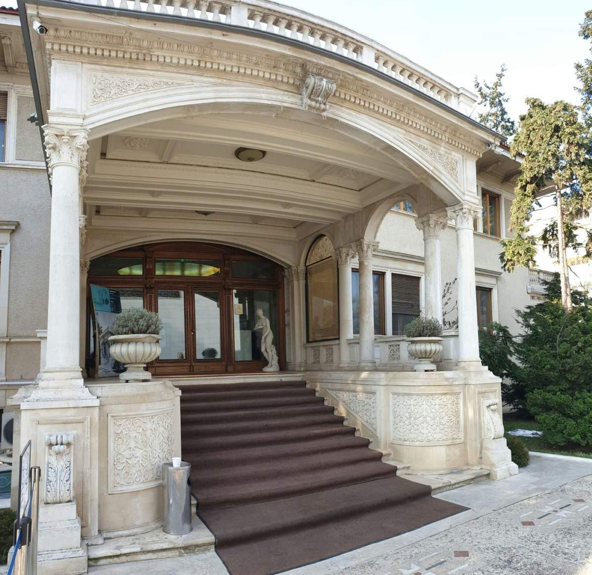 A grand entrance with a set of carpeted stairs leading to wooden double doors, flanked by large planters and ornate stone columns.