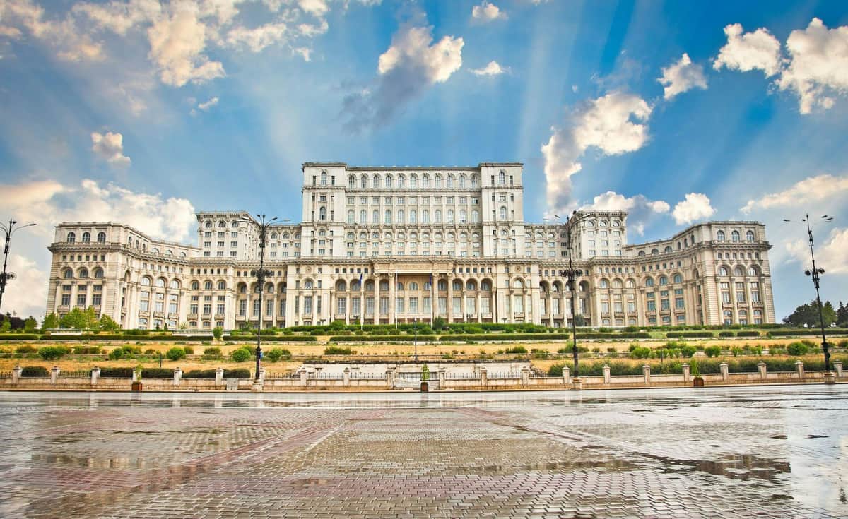 Palace of the Parliament: Entry Ticket + Guided Tour Large neoclassical building with numerous windows and arches, set against a blue sky with clouds, and a wet pavement foreground.