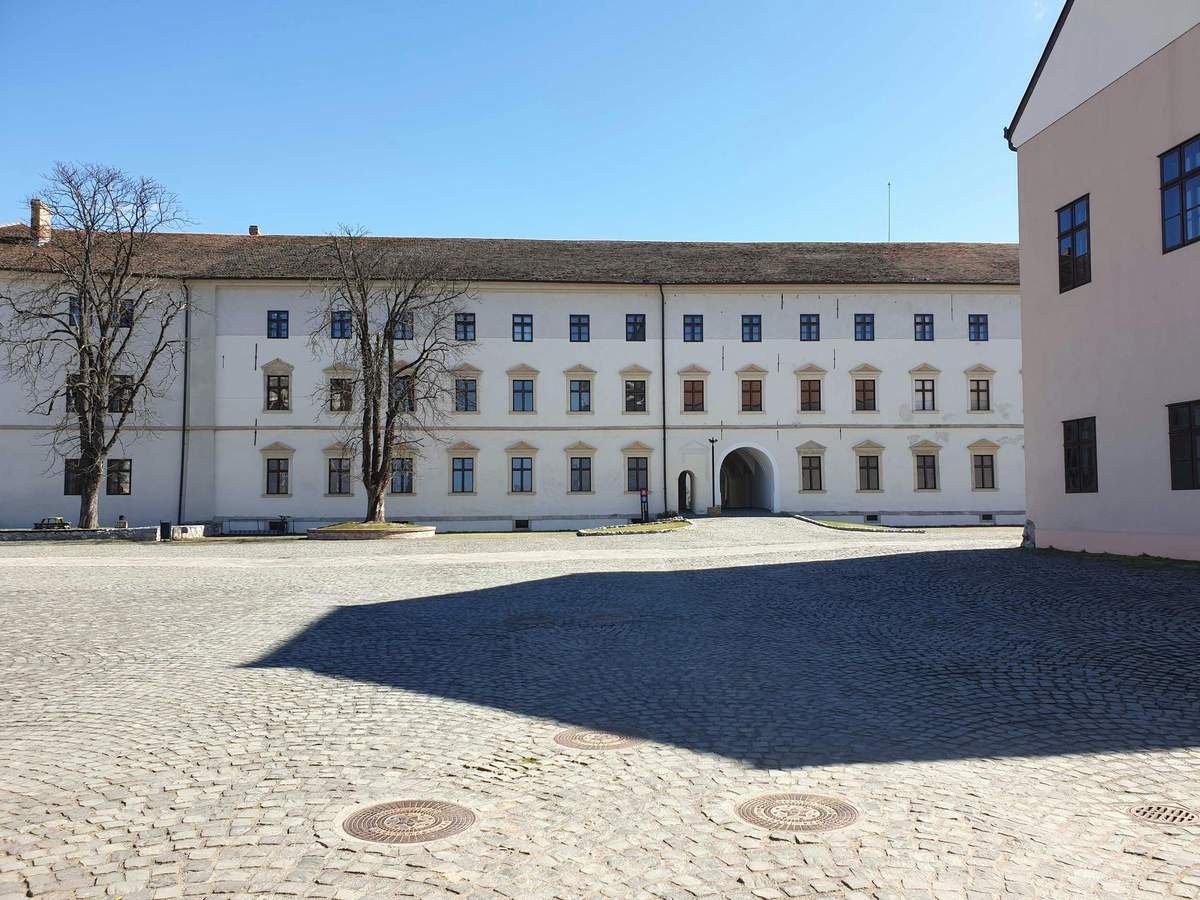 Tour del Museo di Oradea e della Fortezza di Oradea Un grande edificio bianco con diverse finestre e un ingresso ad arco, situato in un cortile di ciottoli sotto un cielo azzurro e limpido.