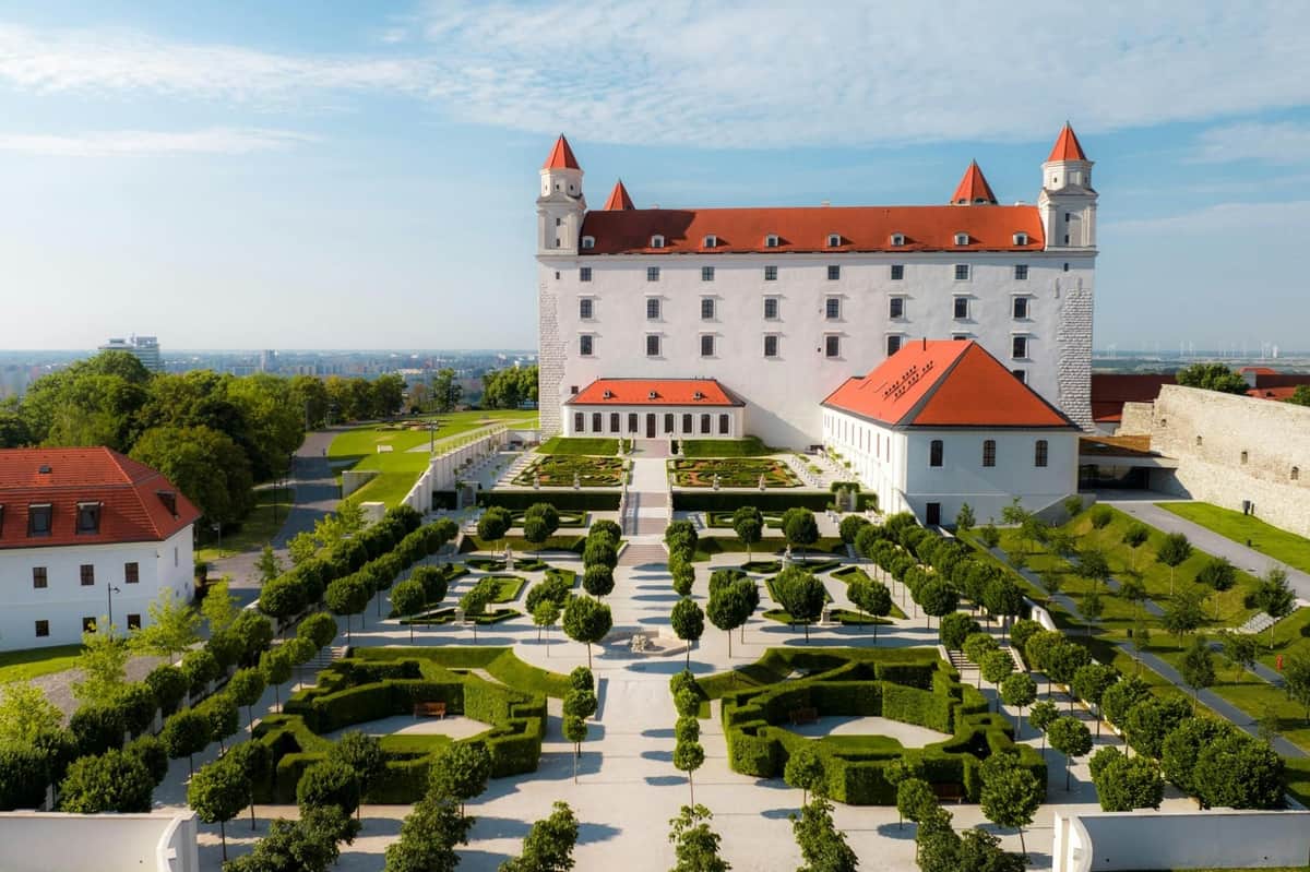 Bratislava: Guided Walking Tour + Castle Entry A castle with a red roof and multiple towers overlooks a manicured garden with geometric hedges and tree-lined pathways.