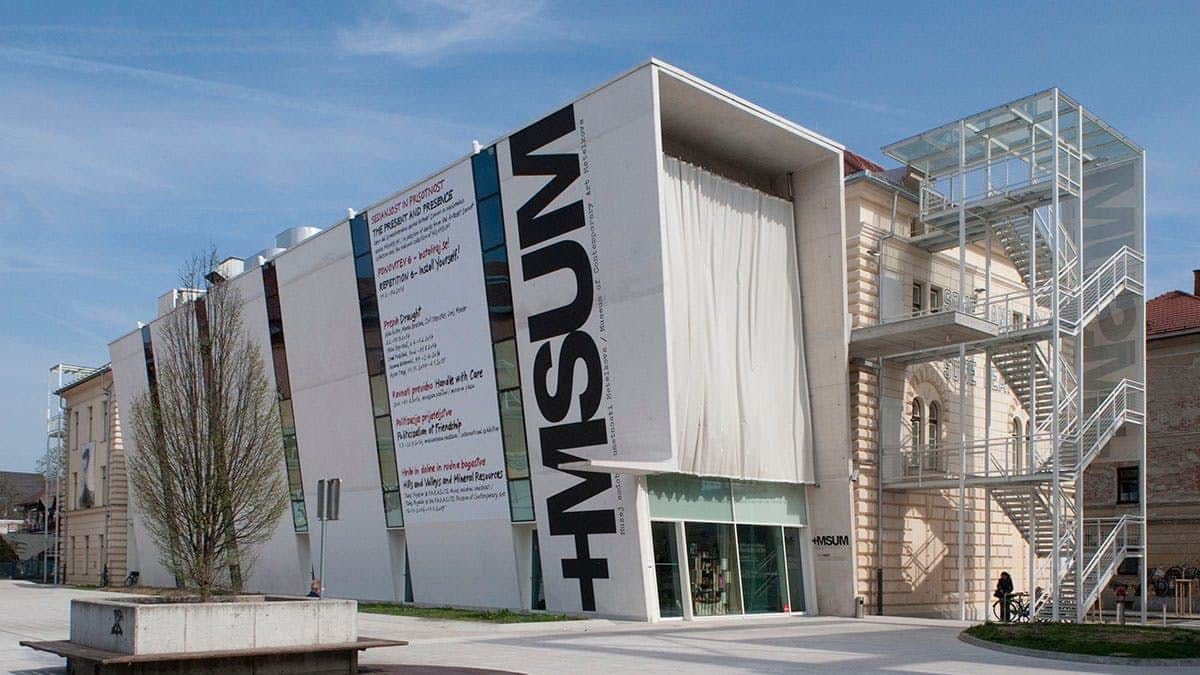 Ljubljana Card Modern building with a unique, tilted design and visible signage saying 'MSUM' on the facade, under a clear sky.