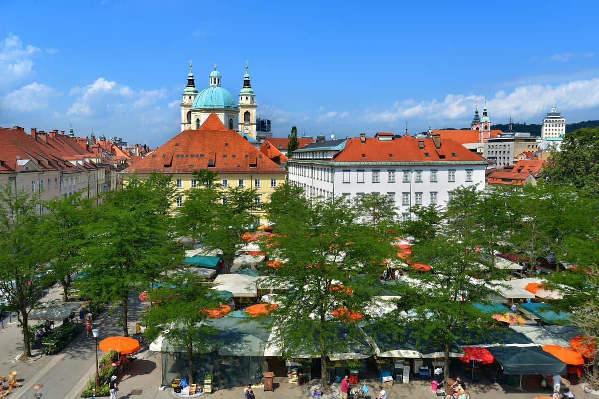 An outdoor market with stalls beneath trees, set in a plaza surrounded by colorful buildings and a prominent church with green domes.