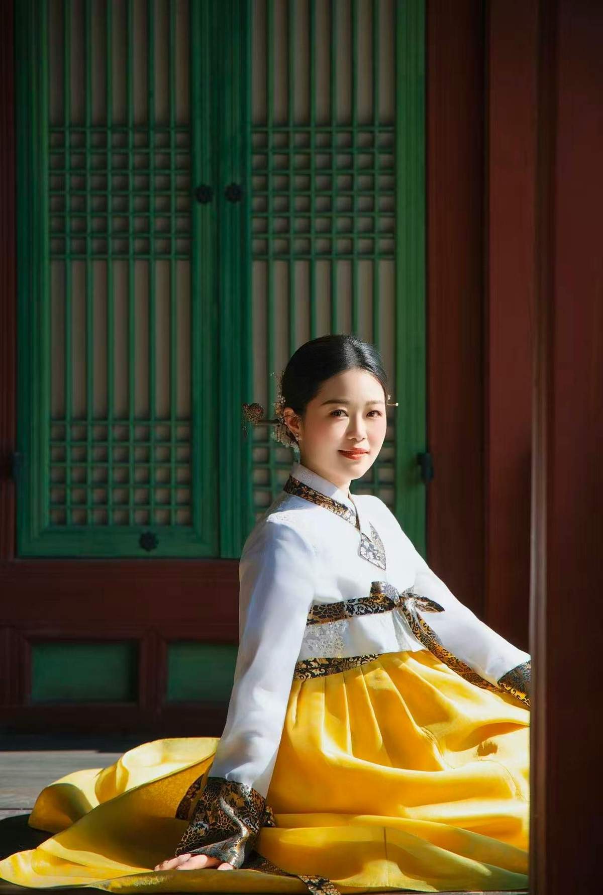 Uma mulher vestida com o traje tradicional coreano, o hanbok, posa em frente a uma porta de madeira com painéis decorativos verdes.