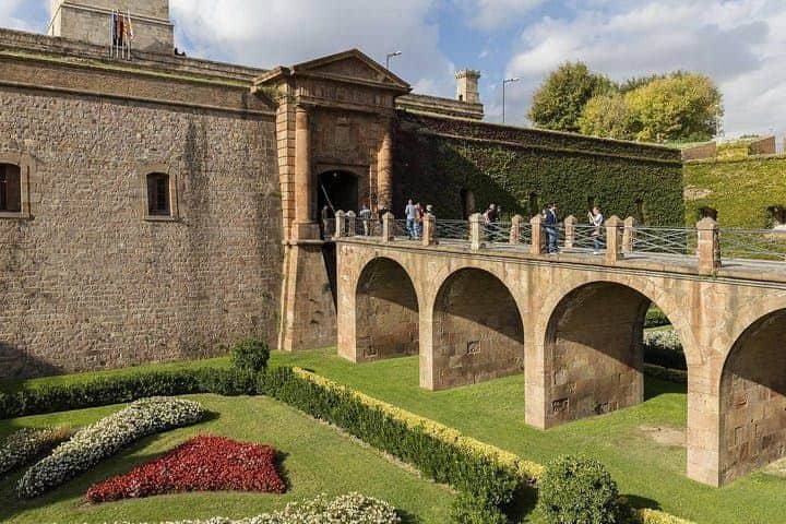Stone bridge leading to an ancient fortress entrance, with people walking. Manicured gardens with colorful flowers are below.