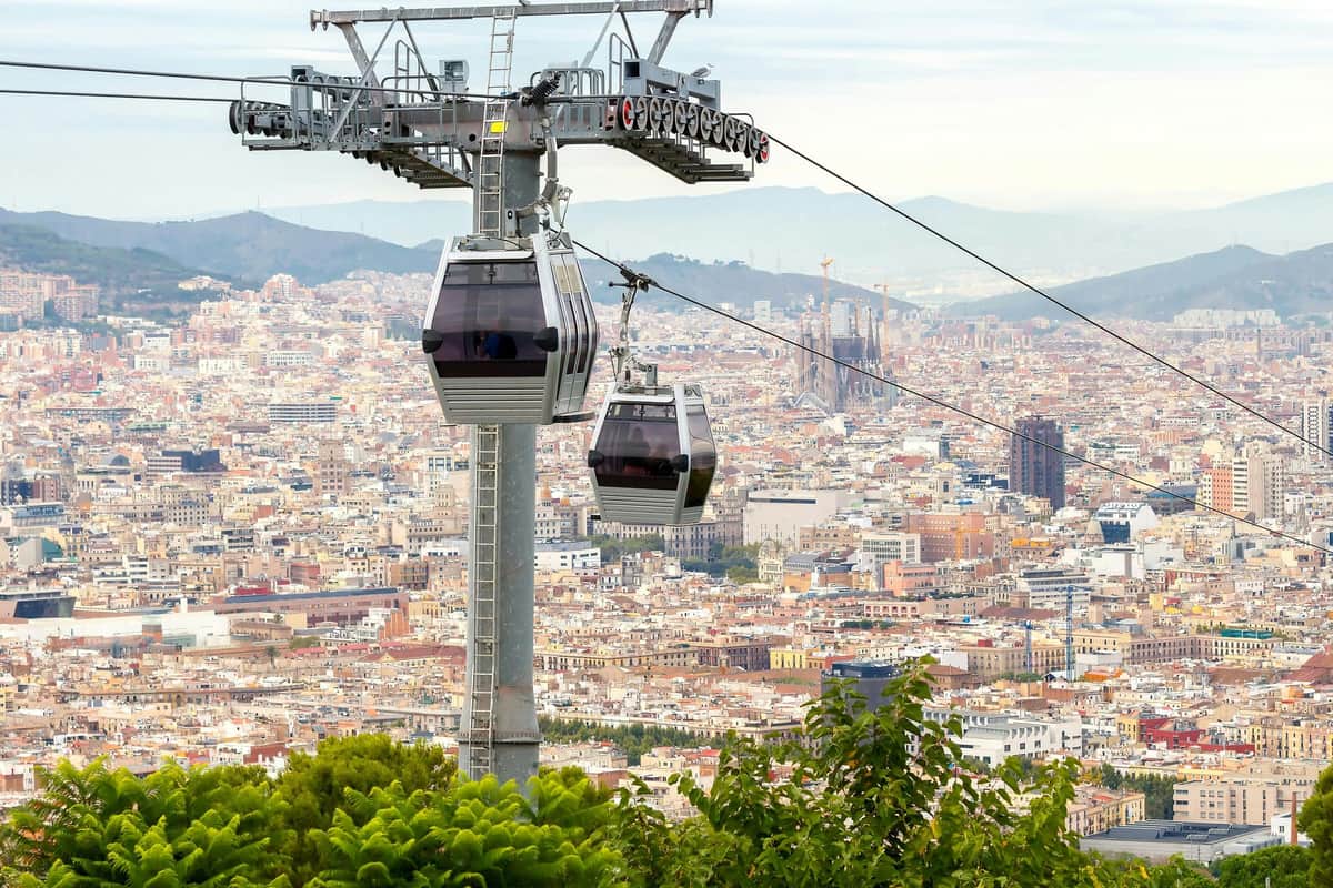 Two cable cars travel over a densely built city with mountains in the background and greenery in the foreground.