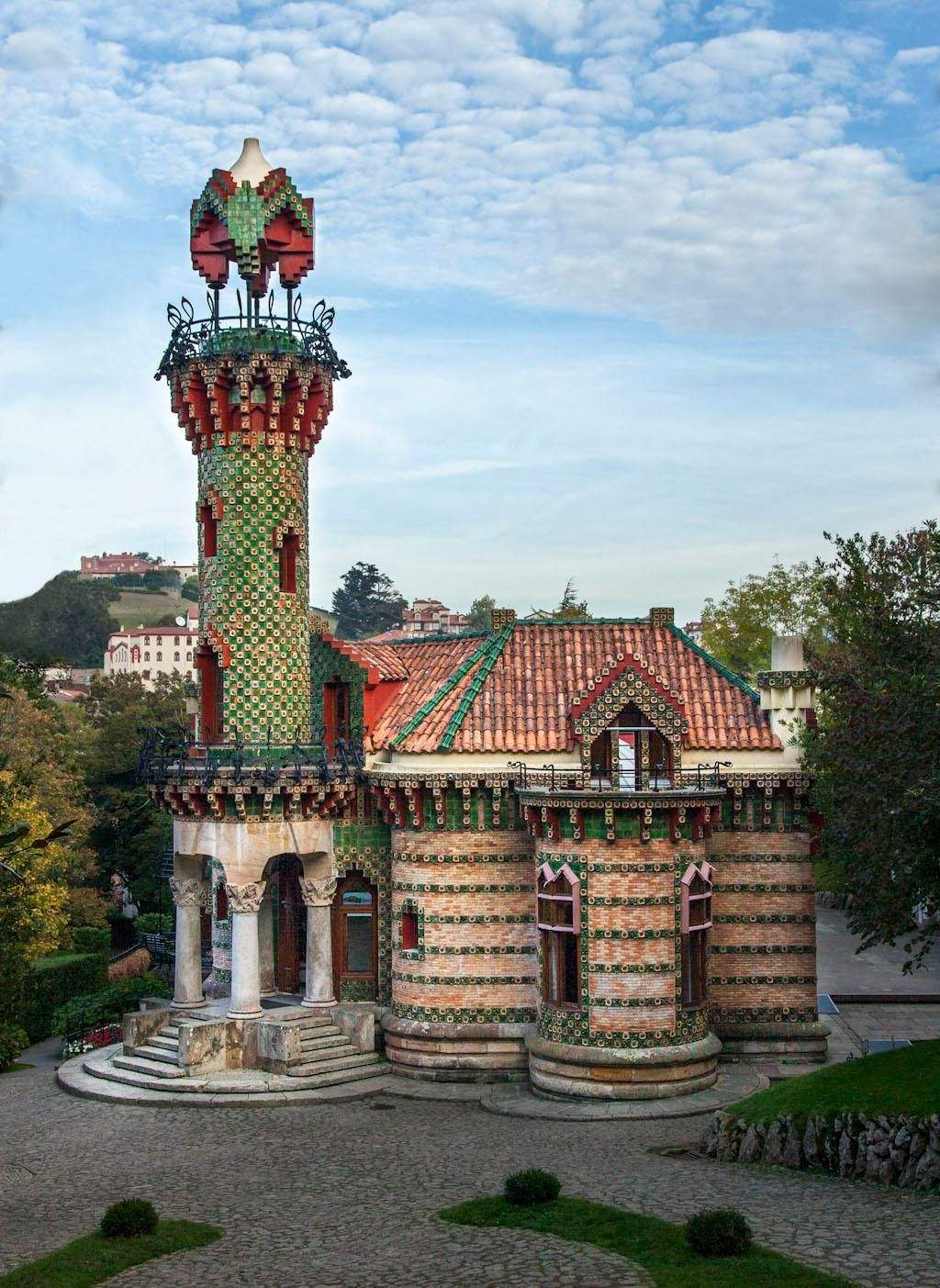 A decorative building with a red-tiled roof and a mosaic-covered tower, surrounded by greenery on a sunny day.