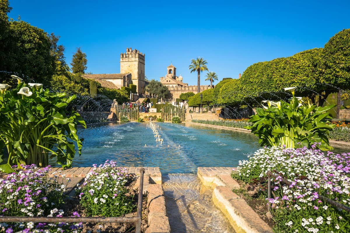 Alcázar des Rois Chrétiens : visite guidée Une grande fontaine entourée de fleurs et de verdure, avec des bâtiments historiques et un clocher en arrière-plan.