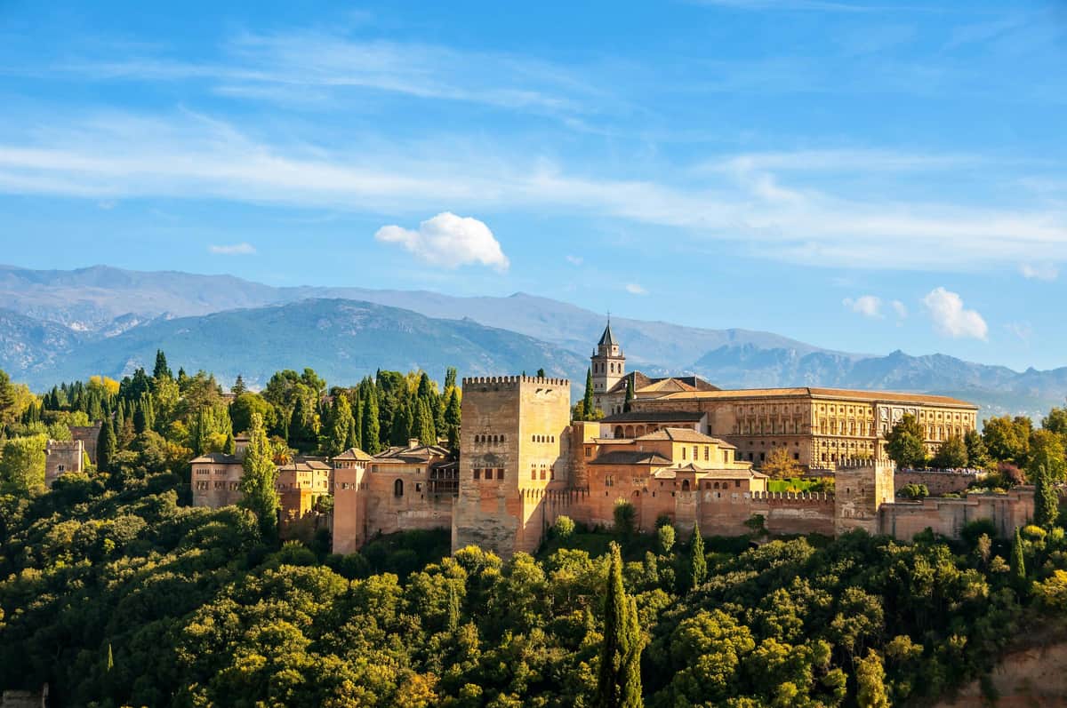 A historic fortress with stone towers and buildings is surrounded by lush greenery, with mountains in the background under a blue sky.