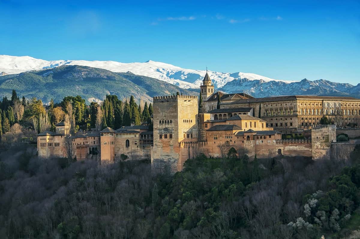 A historic fortress with tower and buildings on a wooded hill, set against a backdrop of snow-capped mountains and a clear blue sky.