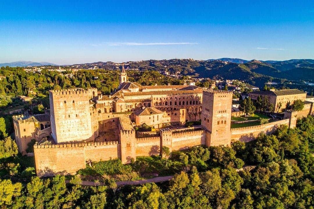 Alhambra: Geführte Tagestour + City Pass Ein historischer Festungskomplex auf einem Hügel, umgeben von üppigem Grün, mit einer Stadt und Bergen im Hintergrund unter einem klaren Himmel.