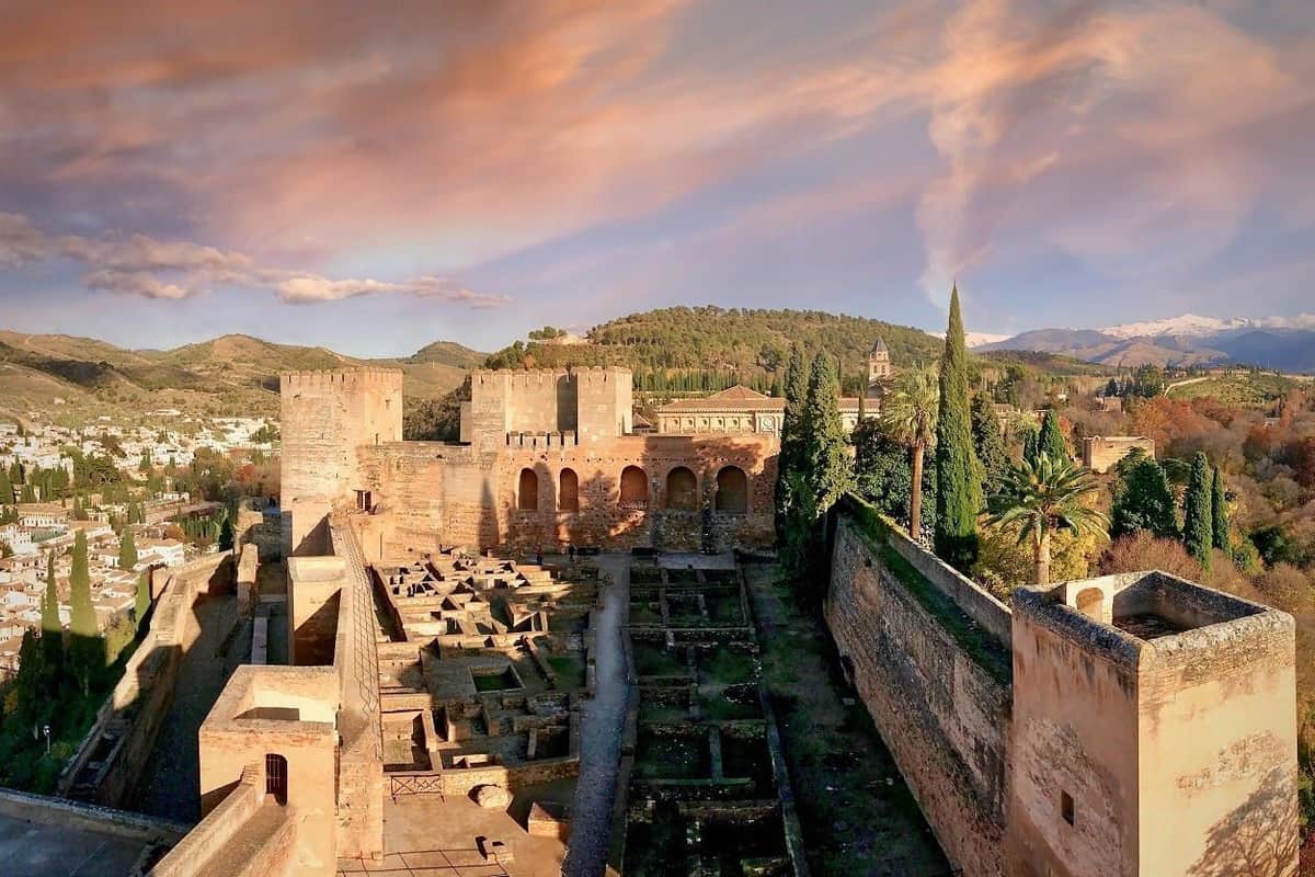 Alhambra, Nasrid Palaces, Albaicín & Sacromonte: Private Guided Tour A historical fortress with stone walls and towers surrounded by greenery under a colorful sky at sunset.
