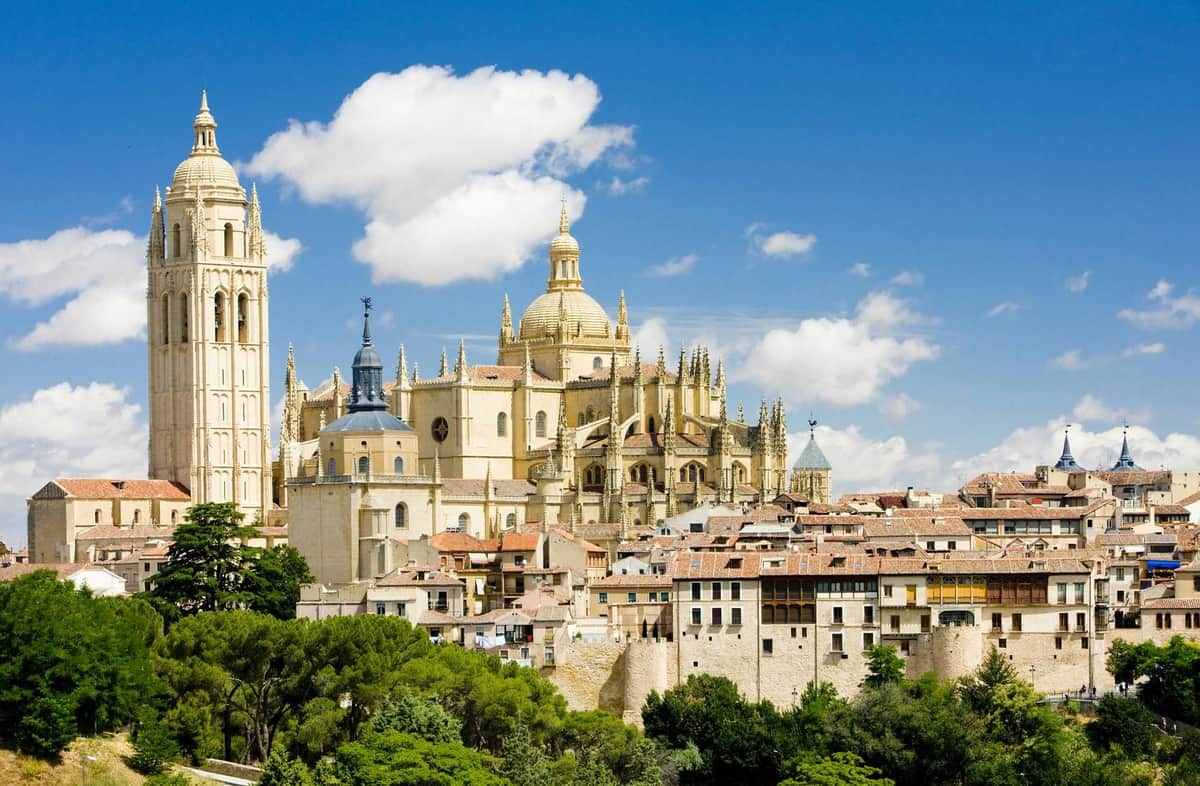 Ávila & Segovia: Day Trip from Madrid + Entry to Alcázar of Segovia (Optional) View of a Gothic cathedral with spires and large central dome, surrounded by historic buildings and lush greenery under a blue sky.