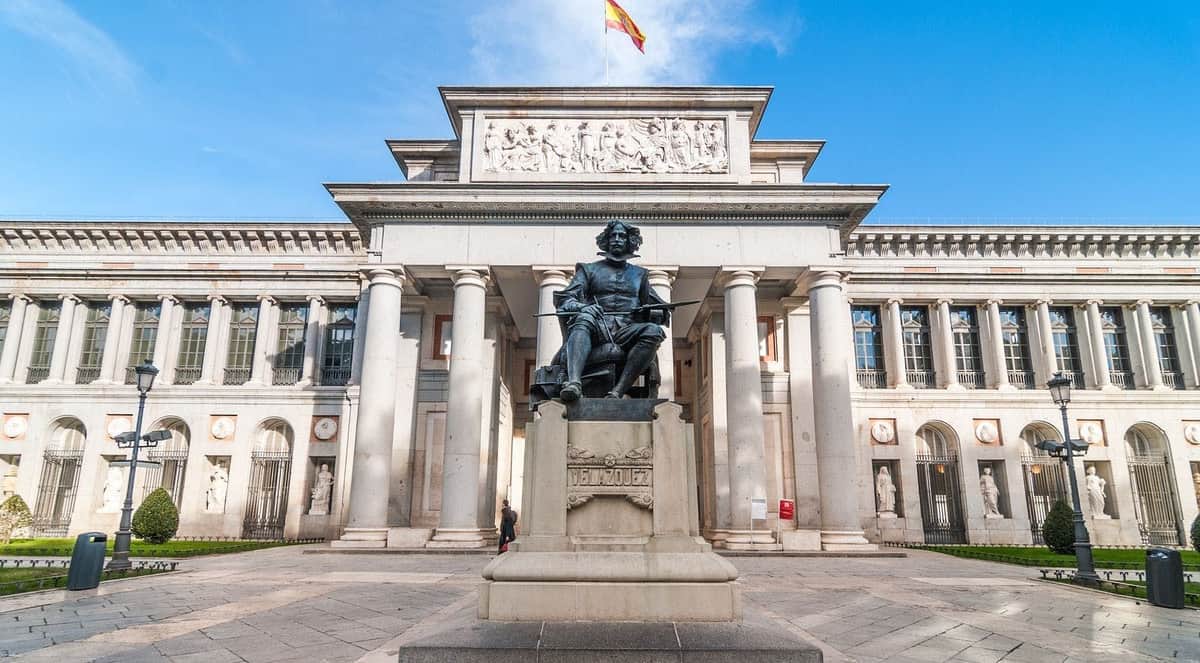Prado Museum & Royal Palace of Madrid: Skip the Line & Guided Tour Statue of Diego Velázquez in front of the Prado Museum's columns, with a Spanish flag flying above.